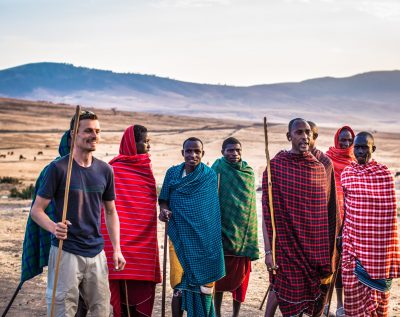 A White male is surrounded by a group of eight African men wearing brightly colored blankets. They are standing in a desert.