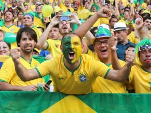 Crowd of soccer fans wearing team colors of yellow and gold. A man in the center has half his face painted green and half his face painted yellow. He is yelling and cheering.