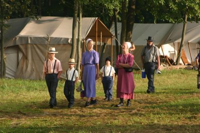 An Amish family are walking outdoors. There are small houses in the distance behind them.