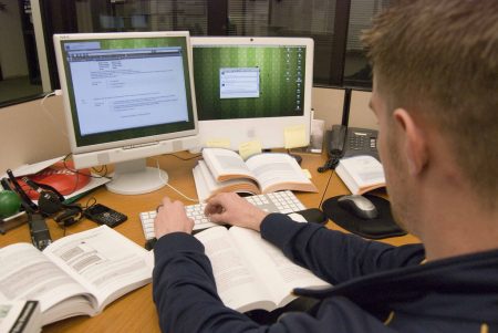 A male student is sitting in front a two computers. There are books and paper in front of the student.